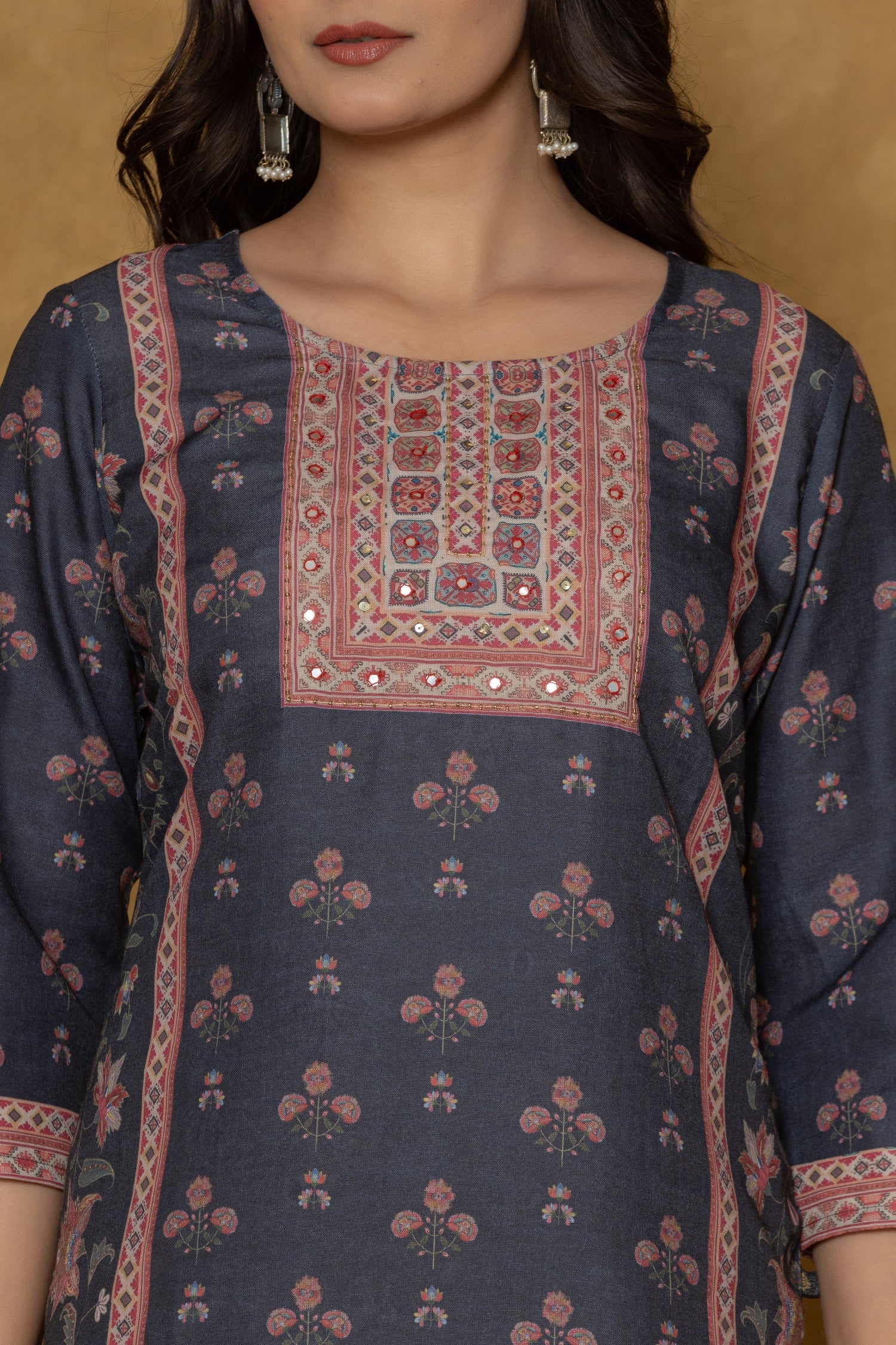 A woman posing in a black kurta detailed with handwork on the round neck, paired with light-colored pants against a textured brown background.