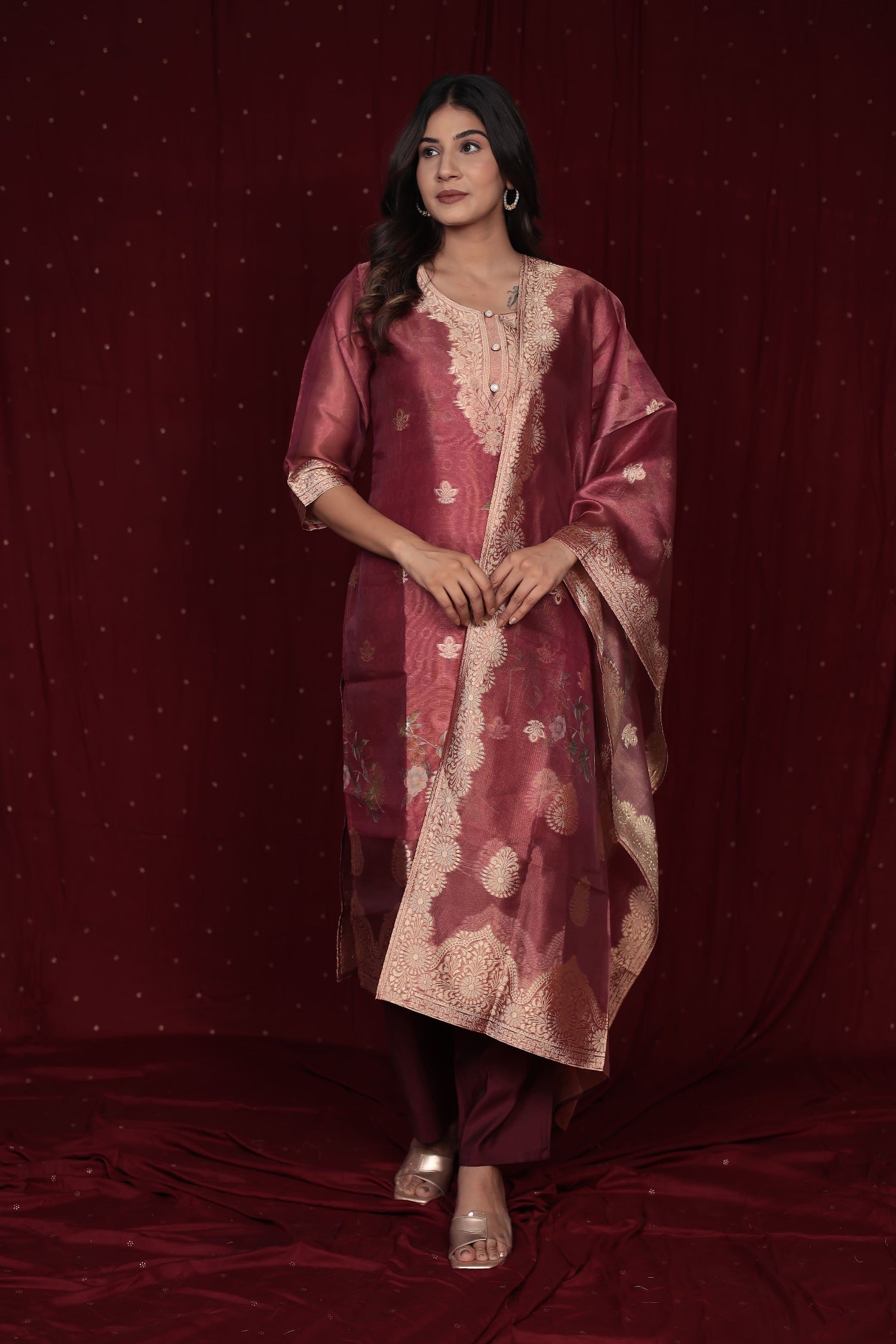 Model wearing a deluxe jacquard organza maroon suit from The Navra, featuring intricate gold woven patterns, embroidered kurti, and a grand jacquard dupatta, with soft silk pants. Perfect for luxurious festive occasions.