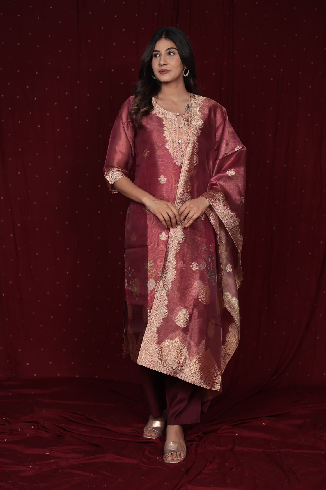 Model wearing a deluxe jacquard organza maroon suit from The Navra, featuring intricate gold woven patterns, embroidered kurti, and a grand jacquard dupatta, with soft silk pants. Perfect for luxurious festive occasions.