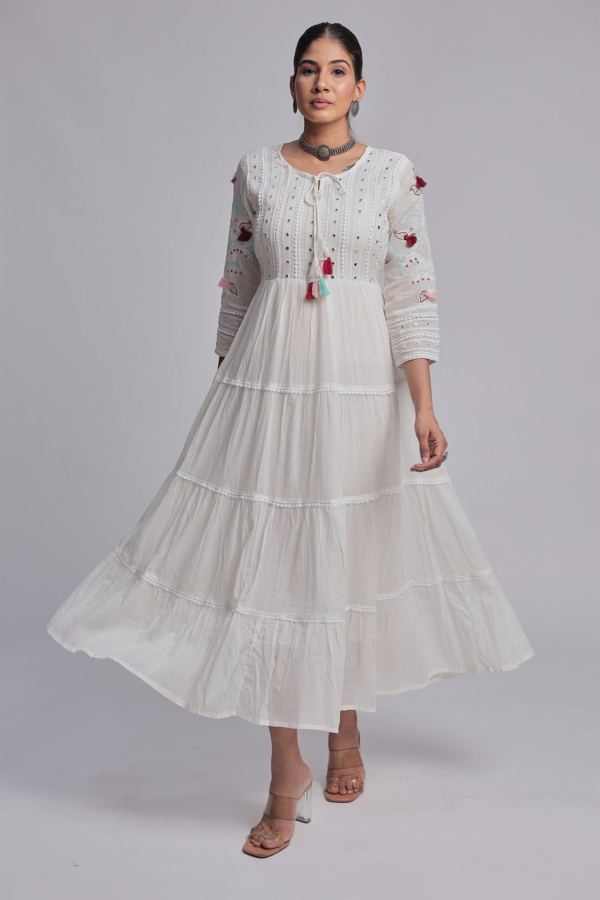 Model wearing a beautiful white tiered Anarkali dress from The Navra, detailed with delicate embroidery, small dark embellishments, and colorful pink, blue, and orange tassels on the front. Ideal for comfortable festive or casual occasions.