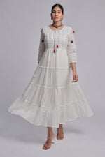 Model wearing a beautiful white tiered Anarkali dress from The Navra, detailed with delicate embroidery, small dark embellishments, and colorful pink, blue, and orange tassels on the front. Ideal for comfortable festive or casual occasions.