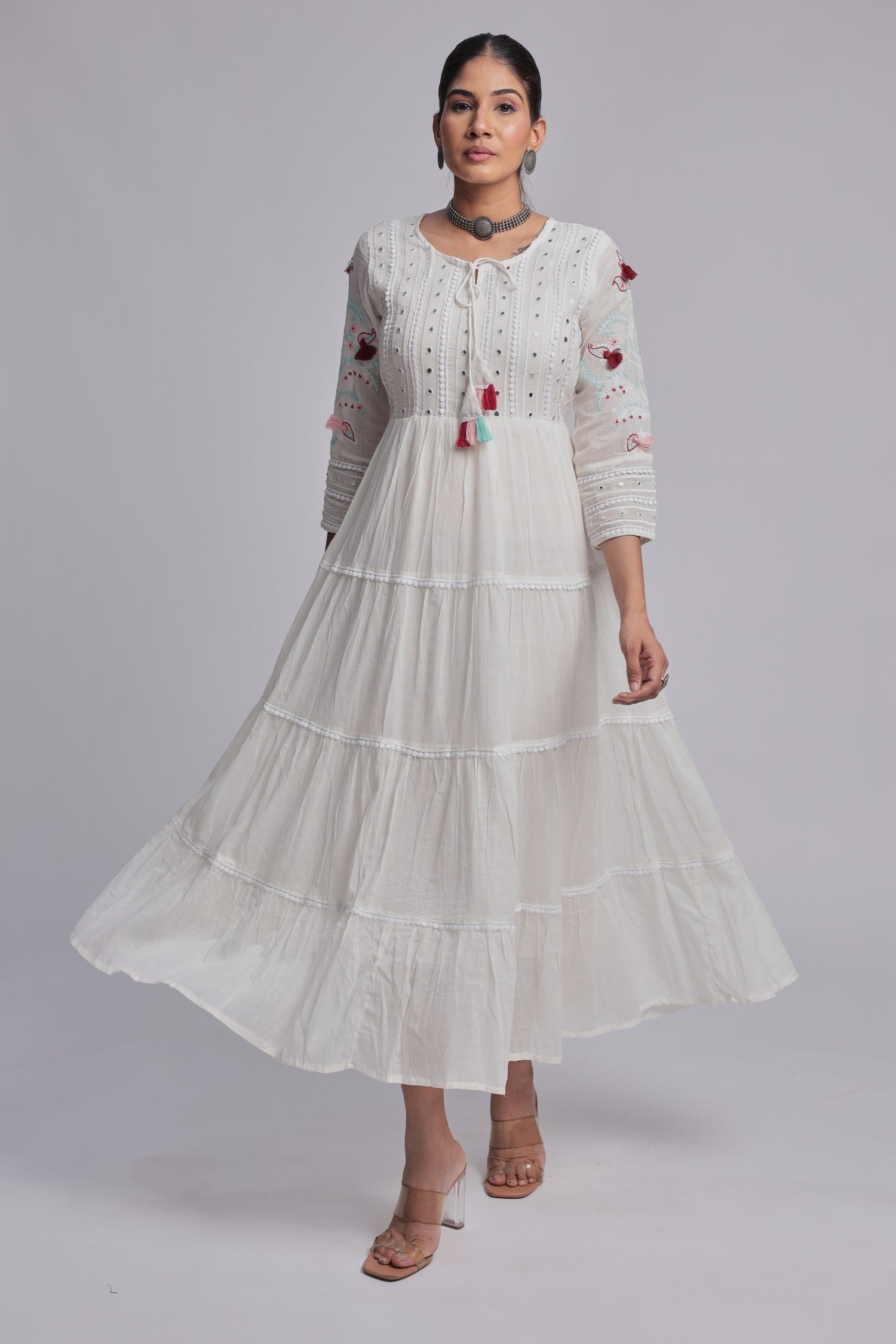 Model wearing a beautiful white tiered Anarkali dress from The Navra, detailed with delicate embroidery, small dark embellishments, and colorful pink, blue, and orange tassels on the front. Ideal for comfortable festive or casual occasions.